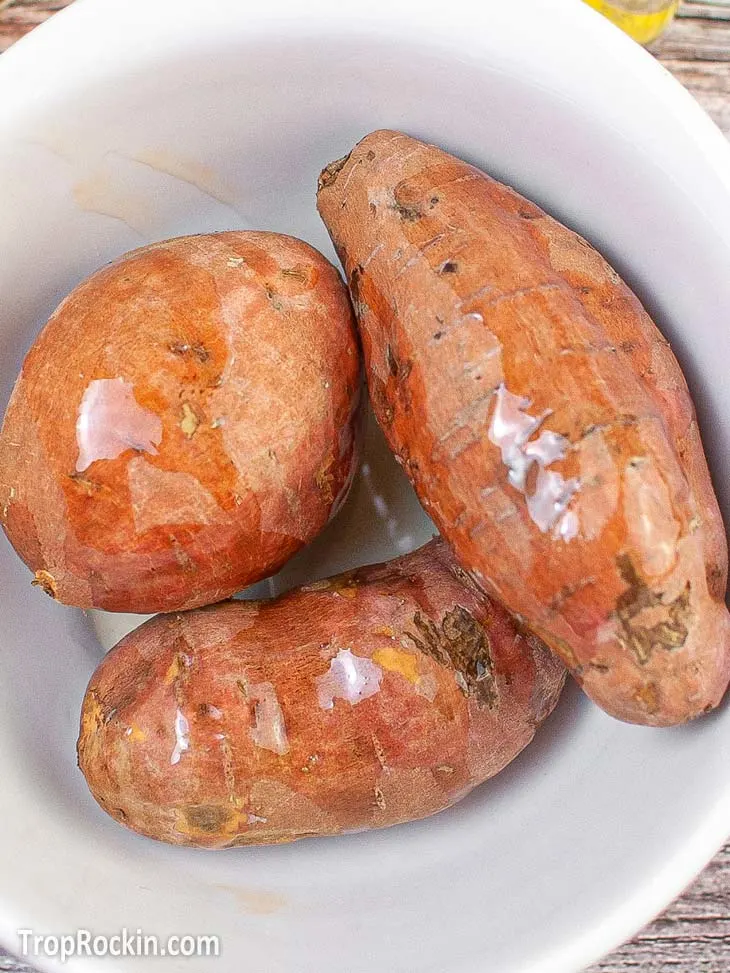 Three medium size sweet potatoes in a white bowl with olive oil on top of them.