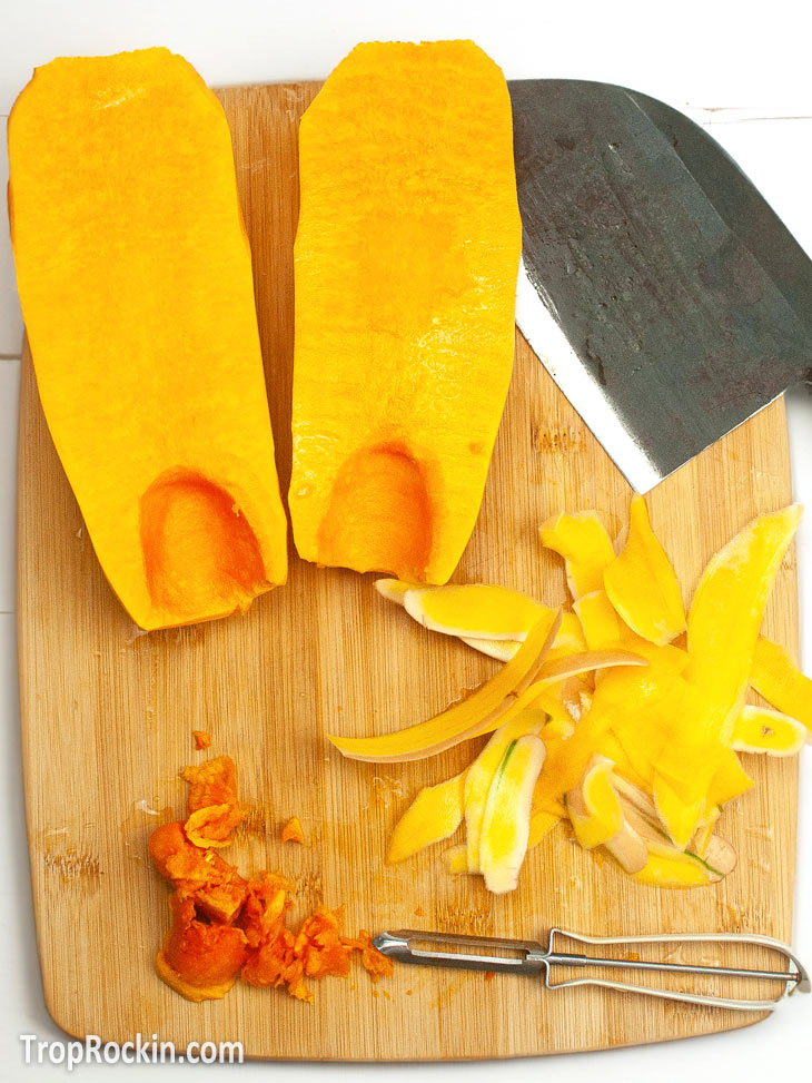 Butternut squash cut in half on a cutting board with a knife and a potato peeler, seeds scooped out and skin peeled. Seeds and skins are also laying on the cutting board.