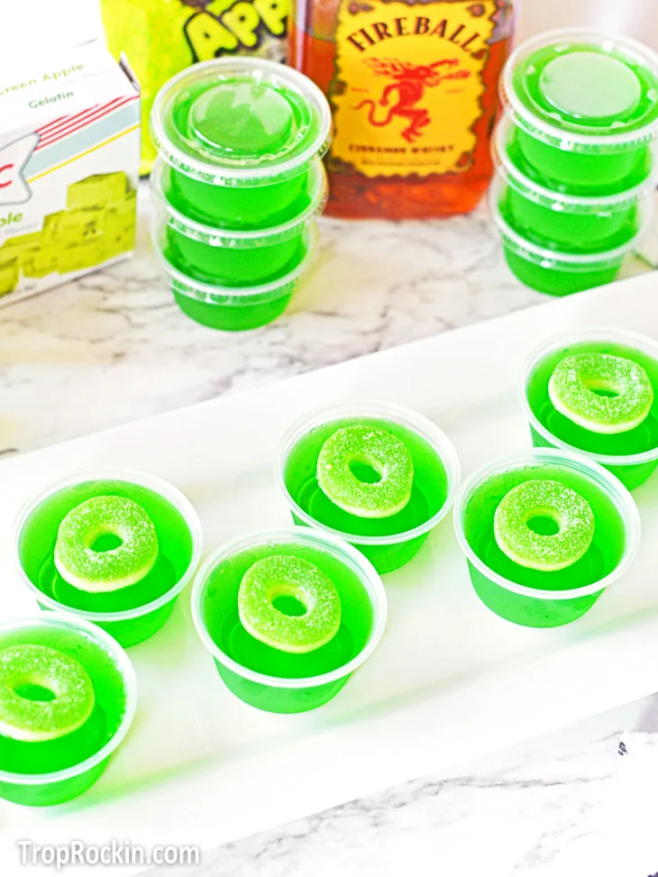 Green apple jello shots lined up on a white rectangular tray. Stacked jello shots and a bottle of Fireball Whiskey in the background.