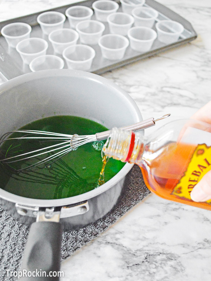 Pouring Fireball Whiskey into a saucepan with green apple jello mixture.