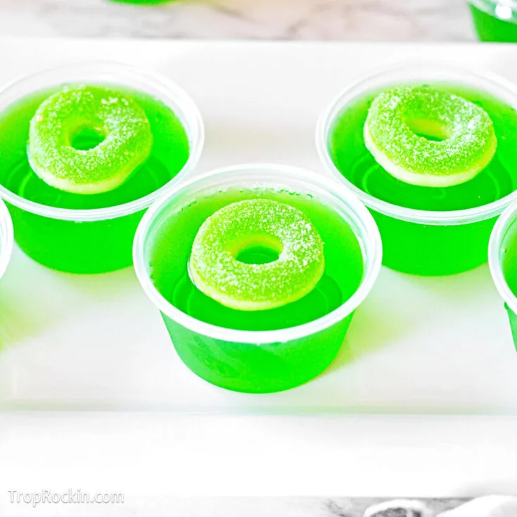 Three green apple jello shots with gummy rings on top sitting on a white serving tray.