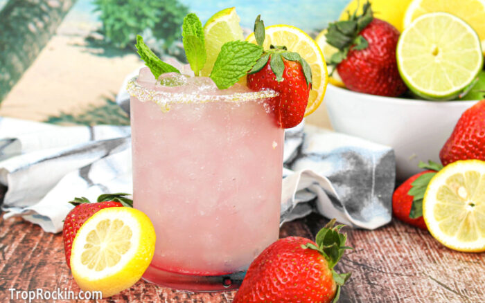 Strawberry Lemonade Margarita in a highball glass on a wooden table with a tropical background. Fresh sliced lemons and whole fresh strawberries decorate the table.