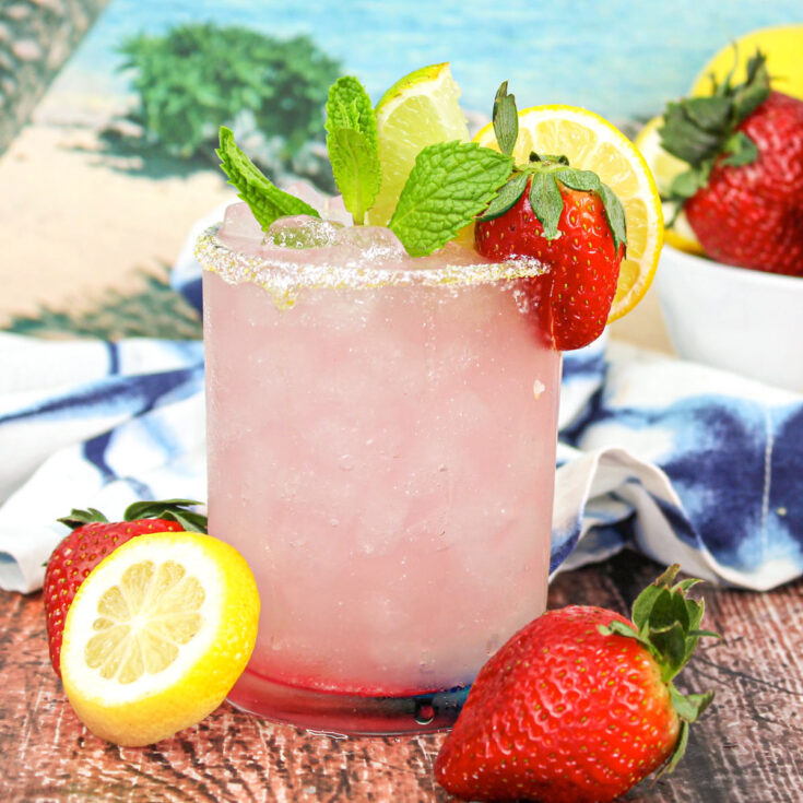 Strawberry Lemonade Margarita with a salted rim, garnished with mint sprig, lemon slice, lime wedge and a fresh strawberry. Sitting on a dark wood table with extra fresh strawberries and lemon slices on the countertop.