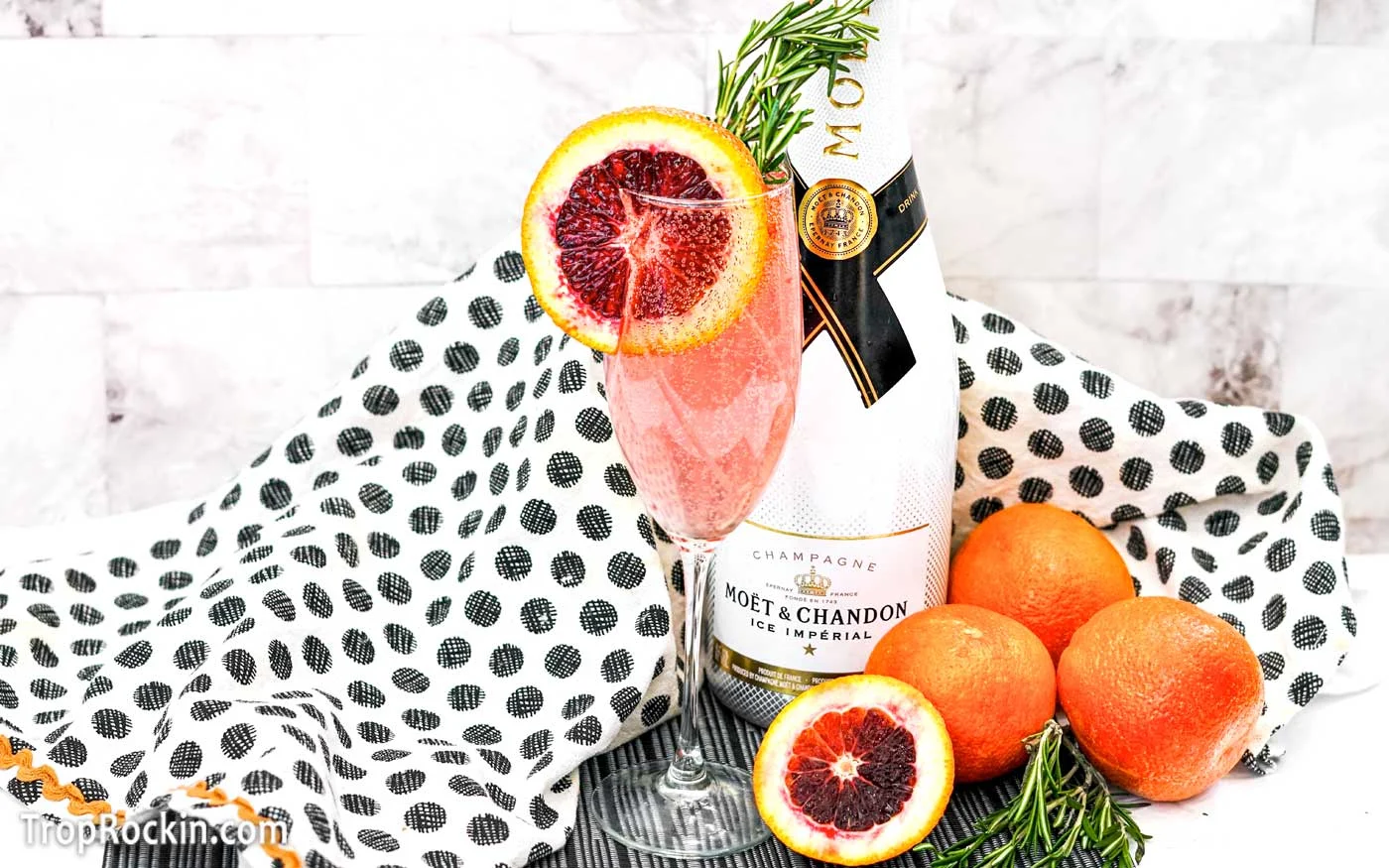 Blood Orange Mimosa with garnishes and a bottle of champagne in the background as well as blood oranges.