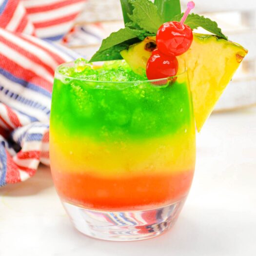 Bob Marley drink with 3 layers: green, yellow and red. Garnished with pineapple slice, maraschino cherries, mint and 2 pineapple leaves. Sitting on a white counter top.