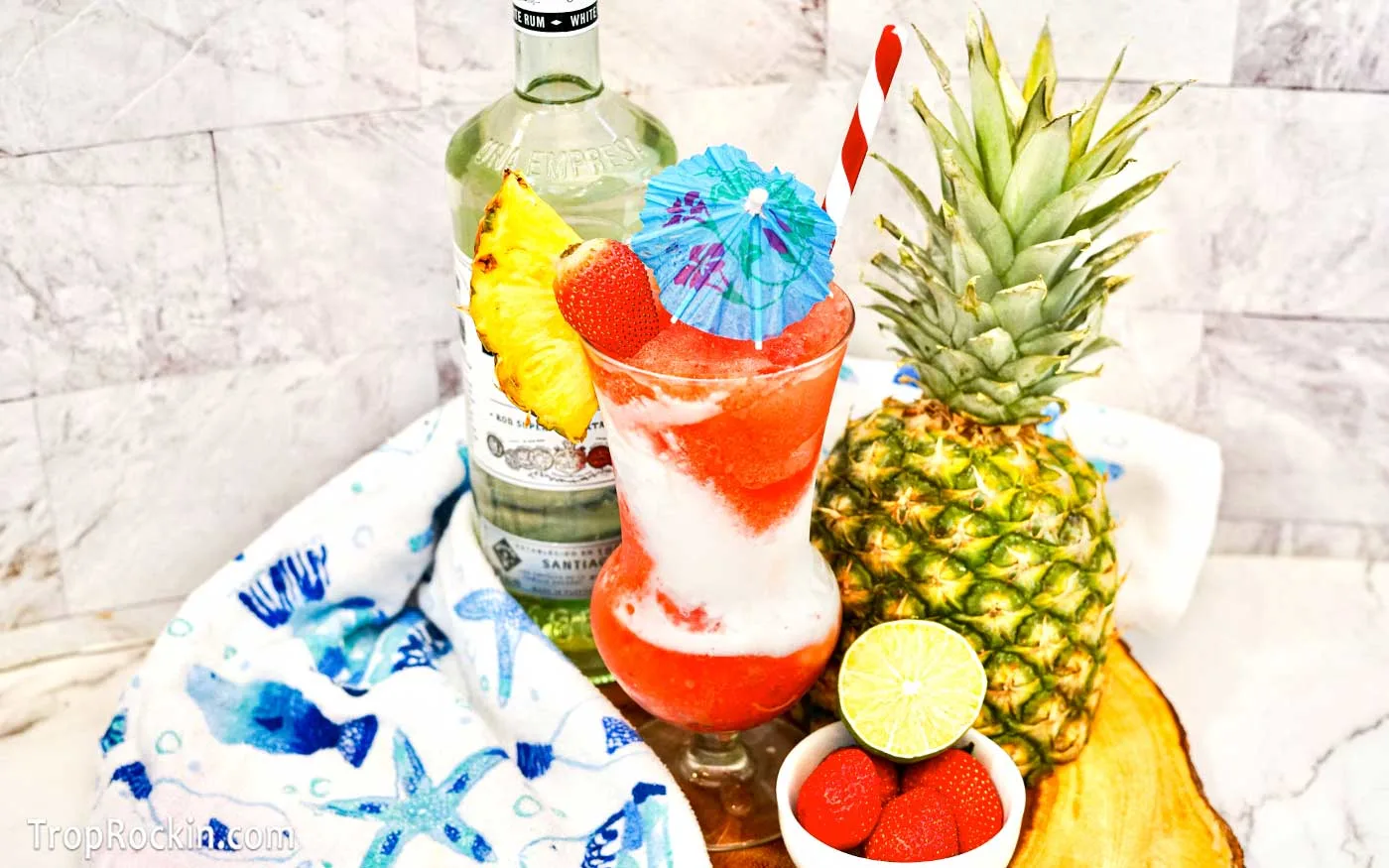 Miami Vice Drink in a hurricane glass with a tropical drink umbrella. Garnished with a slice of pineapple and fresh strawberry. Bottle of Bacardi Light Rum, a fresh pineapple in the background. A bowl of cherries and a half lime in the front.