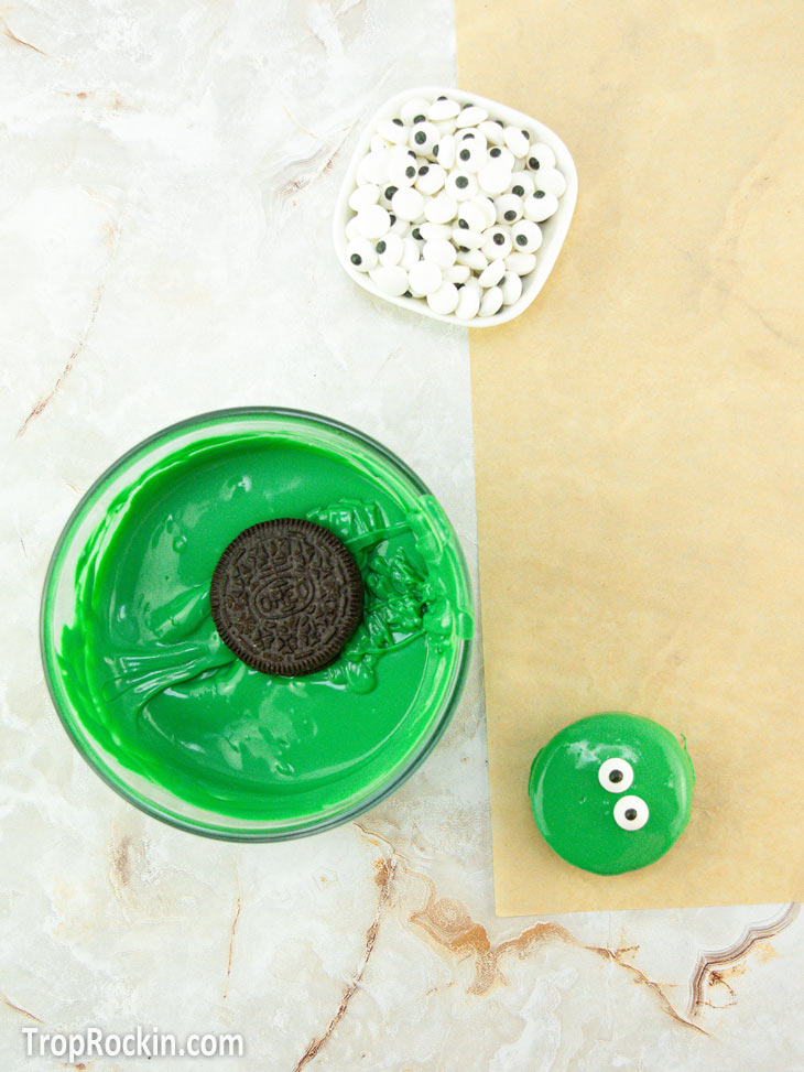 Oreo cookie in a bowl of melted green candy wafers, bowl of candy eyes and a green covered oreo on parchment paper with two candy eyes.
