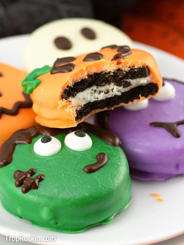 Halloween Chocolate Covered Oreos decorated in four ways: Green Frankenstein, Orange Jack-o-lantern stacked on top with a bite out of the cookie, Purple Vampire and a white ghost.