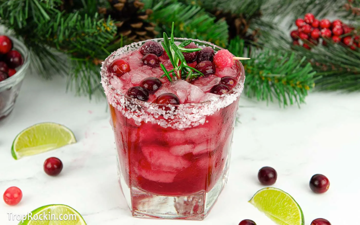 Christmas margarita garnished with cranberries and a rosemary sprig. Limes and cranberries scattered on counter top wih Christmas greenery in the background.