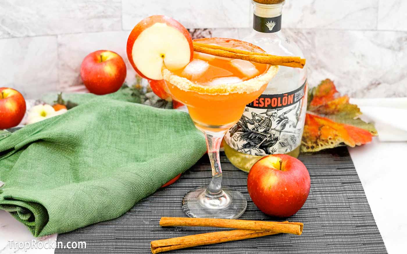 Cranberry Apple Margarita in a margarita glass with brown sugar rim and garnished with an apple wheel slice and a cinnamon stick. A bottle of tequila and whole apples in the background with a green linen napkin.