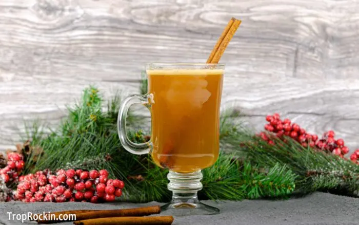 Hot buttered rum drink in a glass mug with a handle and garnished with a cinnamon stick. Christmas greenery behind the drink.