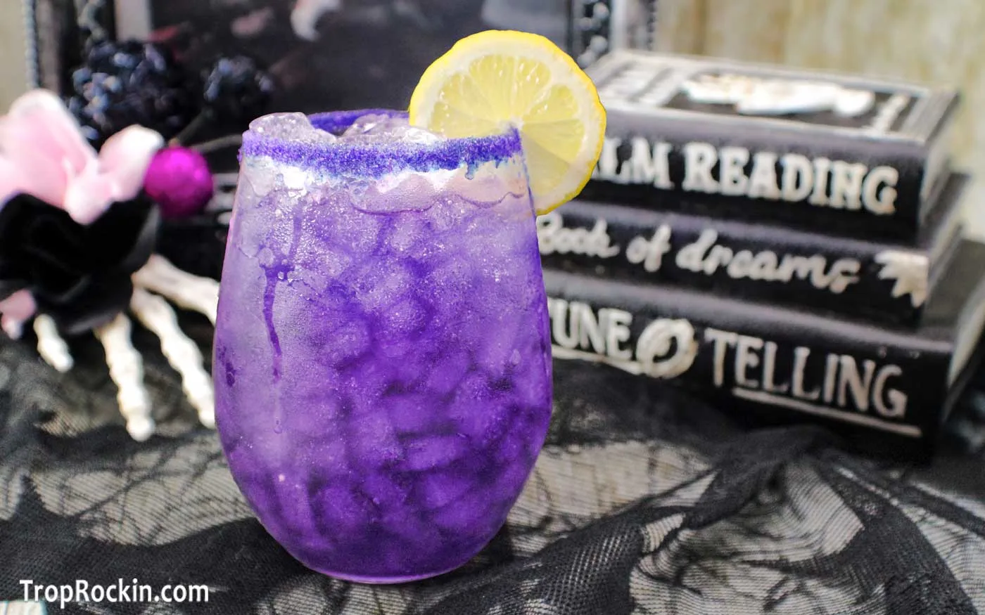 Purple People Eater Drink is a purple color in a cocktail glass with purple sugar on the rim and garnished with a lemon slice. Halloween blurry in the background.