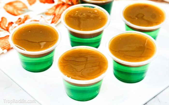 Caramel apple jello shots on white cutting board with fall kitchen towel in background.