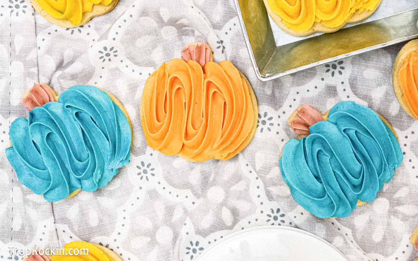 Three pumpkin spice cookies cutout in the shape of pumpkins decorated with a light blue buttercream frosting and one decorated with orange buttercream frosting.