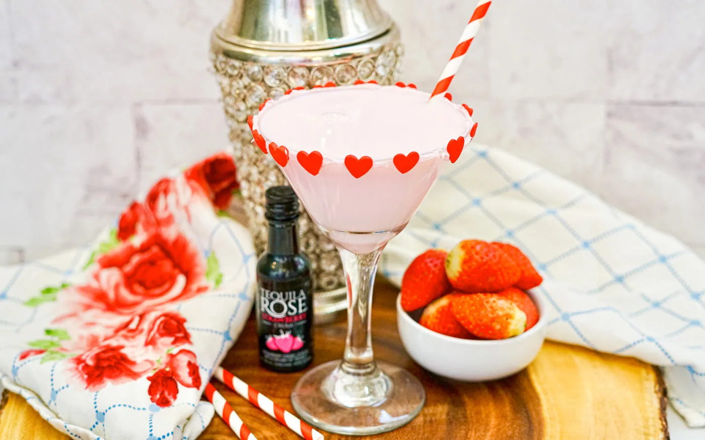 Chocolate strawberry martini with a shaker, bowl of strawberries and a bottle of tequila rose on a table top.