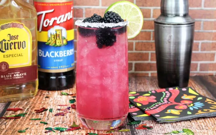 A blackberry margarita with fresh blackberries for garnish and a lime wedge. Tequila bottle, blackberry syrup and a cocktail shaker in the background.