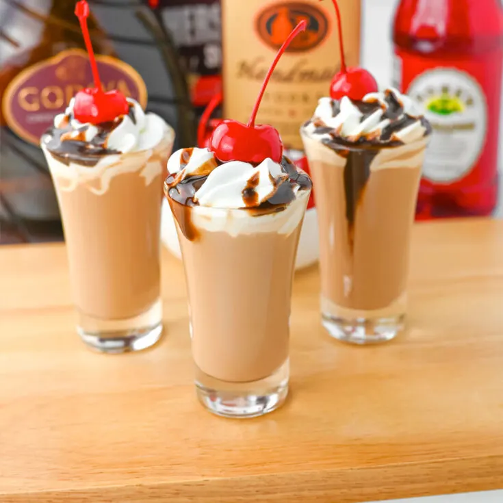 Three chocolate covered cherry shots topped with whipped cream, chocolate sauce and maraschino cherries sitting on a cutting board with liquor bottles in the background.
