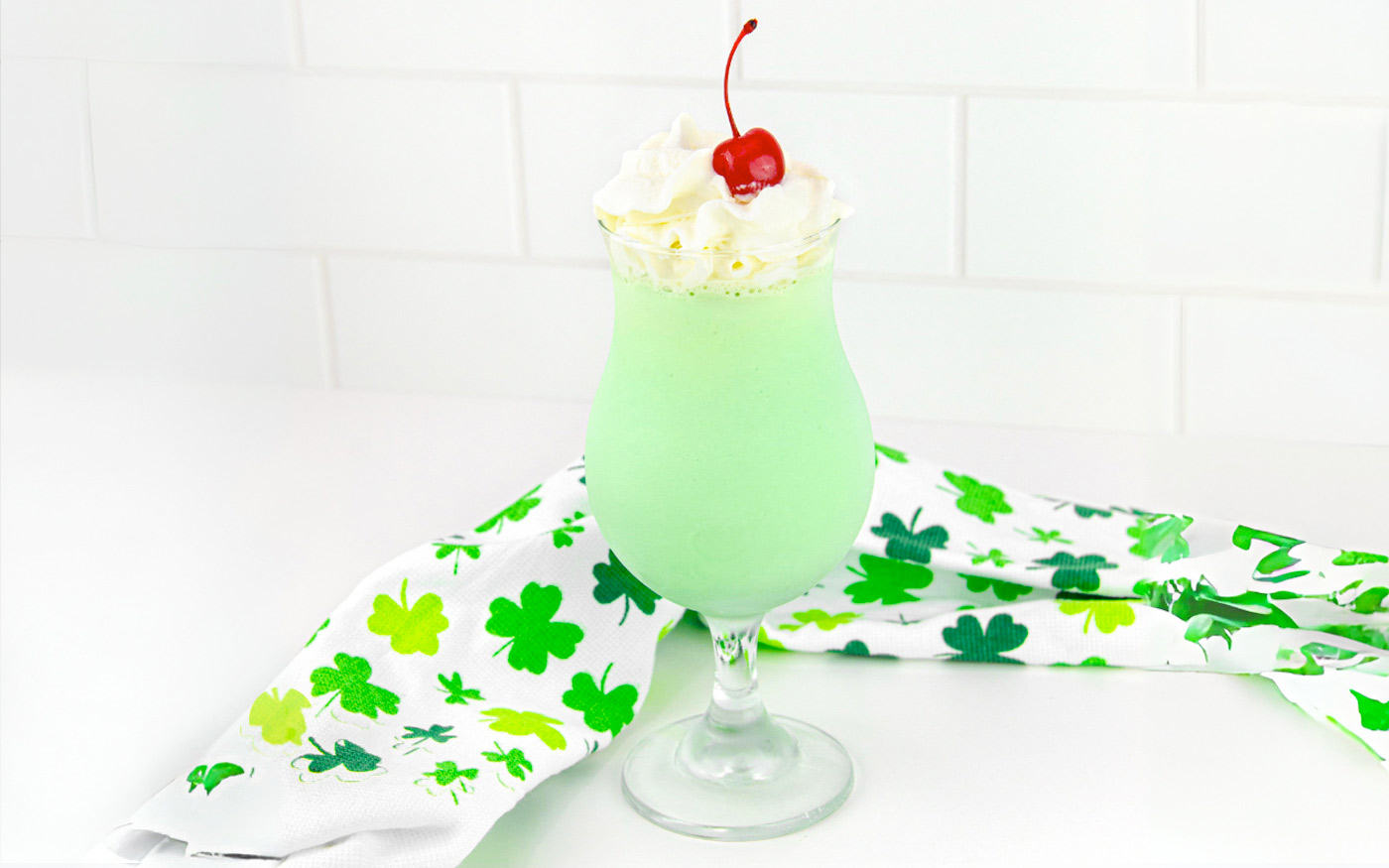 Boozy Shamrock Shake which is a green shake with whipped cream and a maraschino cherry. St. Patrick's day style kitchen towel on counter top.