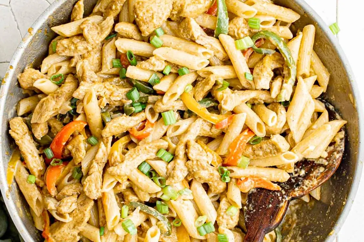 Creamy Jerk chicken pasta with red, green and yellow peppers in a large mixing bowl with a wooden spoon.